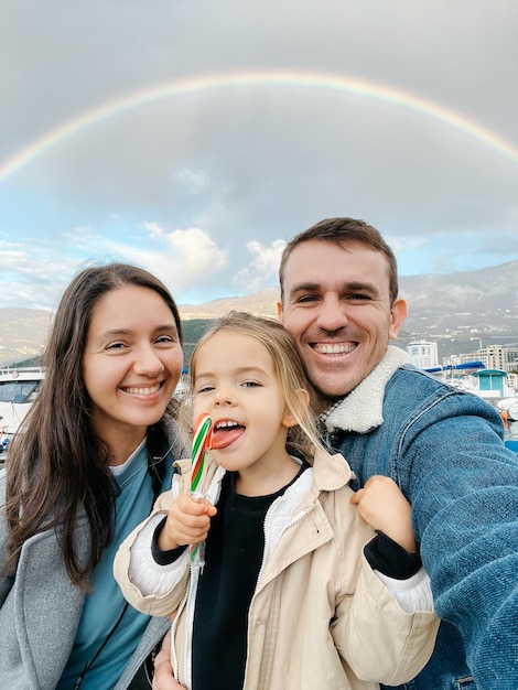 写真 お父さんお母さんと山にかかる虹の背景にロリポップを持つ小さな女の子