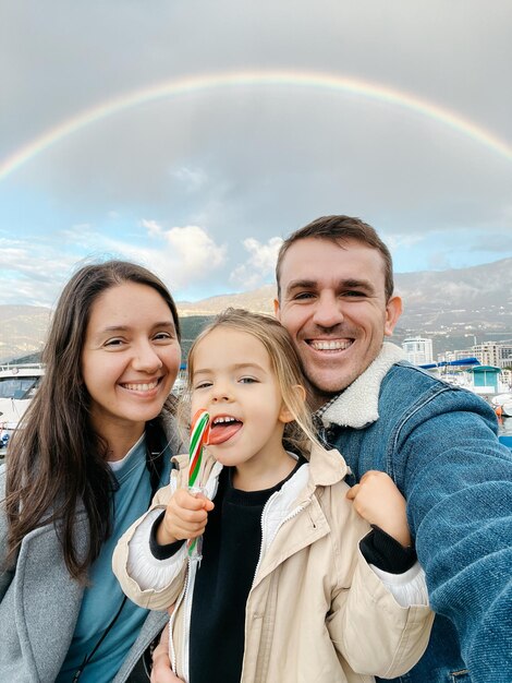 Dad mom and little girl with lollipop on the background of the rainbow over the mountains