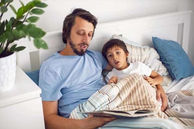 Dad reads a book lying in a white bed to his son child before going to bed