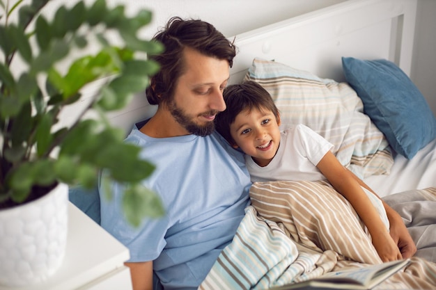 Dad reads a book lying in a white bed to his son child before going to bed