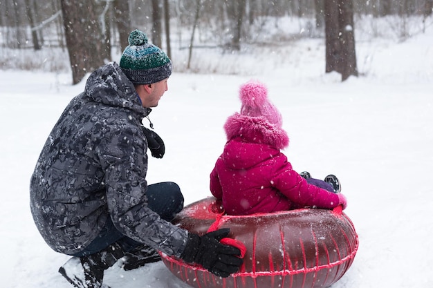 Dad rides his daughter on a tube from a slide in winter in a
snowfall. tubing, winter sports outdoor and family entertainment.
warm clothes, knitted hat with a pompom. help and support, family
values