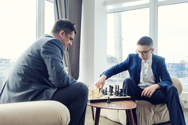 Dad and son are sitting at a table by a wide window and playing chess making moves with pieces