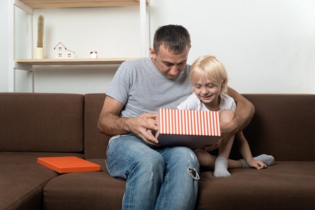 Dad and son open gift box while sitting on the couch at home.