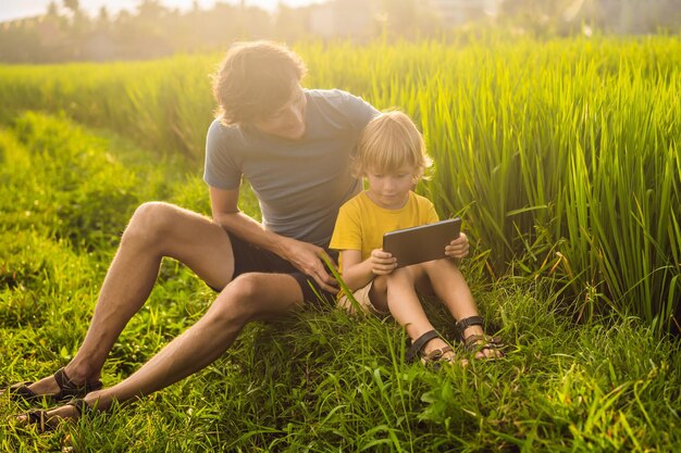 태블릿을 들고 들판에 앉아 있는 아빠와 아들 화창한 날 잔디에 앉아 있는 소년 홈 스쿨링 또는 태블릿 게임