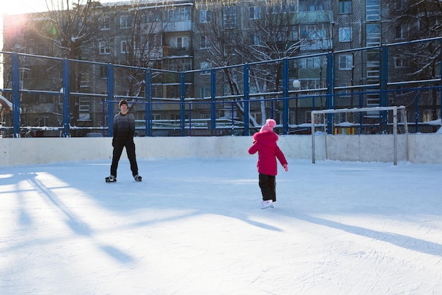 写真 お父さんは幼い娘に、市内の高層ビルの中庭にあるスケートリンクでアイススケートをするように教えています。凍るような冬の晴れた日、アクティブなウィンタースポーツとライフスタイル