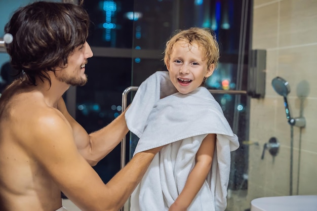 Dad wipes his son with a towel after a shower in the evening before going to sleep on the background of a window with a panoramic view of the city