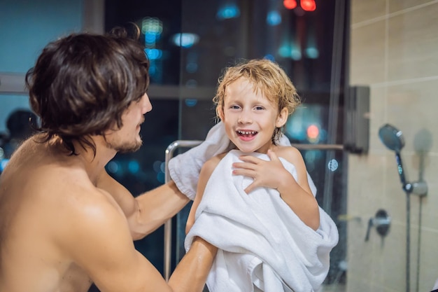 Dad wipes his son with a towel after a shower in the evening before going to sleep on the background of a window with a panoramic view of the city