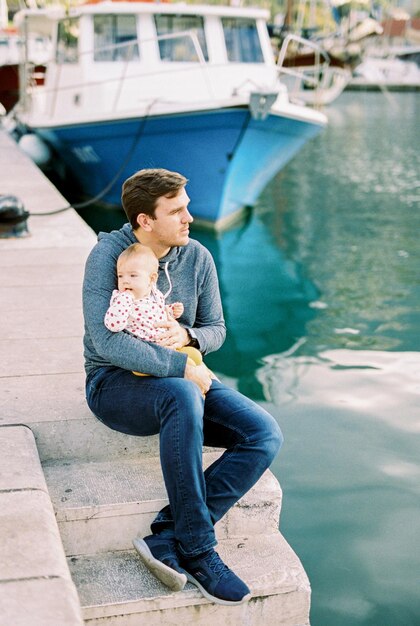 Dad with a baby on his knees sits on the pier near the boat and looks at the sea