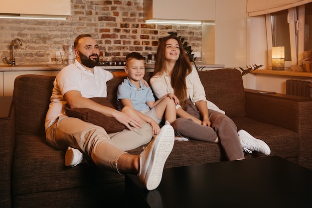 Dad with a beard, son, and young mom with long hair are watching with interest TV and smiling on the sofa in the apartment