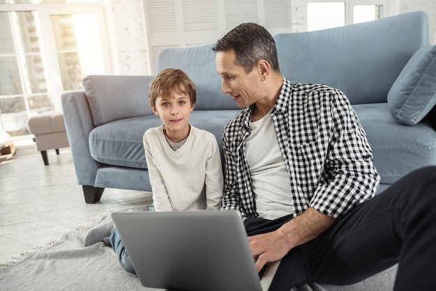 Dag samen. Knappe waakzame blonde jongen glimlachend en zittend op de vloer met zijn vader en zijn vader met een laptop en kijken naar zijn zoon