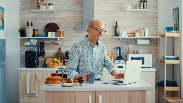 Dagelijks leven van senior man in de keuken tijdens het ontbijt met behulp van laptop met een kopje koffie. Oudere gepensioneerde die vanuit huis werkt, telewerken met behulp van externe internetbaan online communicatie op mod