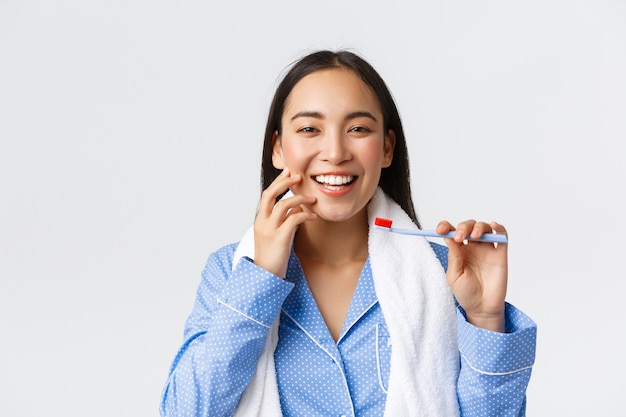 Daily routine, morning and hygiene concept. Close-up of gorgeous asian girl with perfect white smile and clean skin holding towel and toothbrush, brushing teeth in blue pajama, white wall