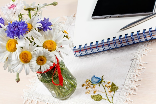 Photo daisy chamomile and cornflowers in vase on a wooden table with a smart-phone, exercise book and a pen