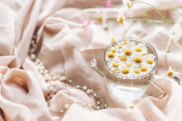 写真 野生の花と宝石と柔らかいベージュの生地の背景にガラスのカップに水にデイジーの花 優しい花の美学 テキスト ボヘミアンな気分のためのスペースを持つ創造的な夏のイメージ