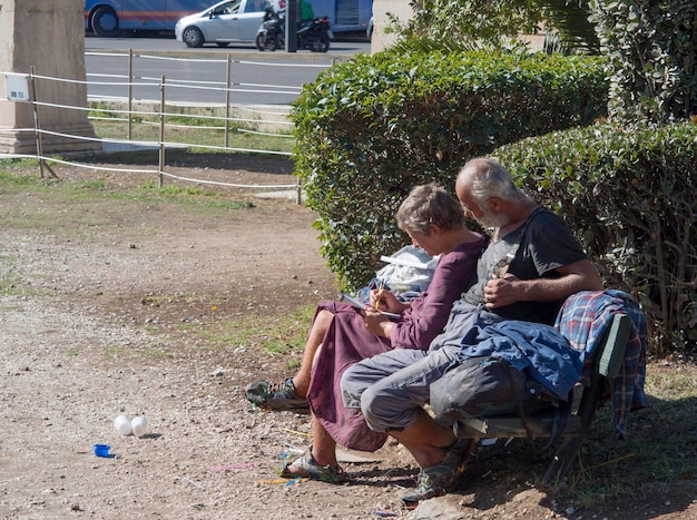 Daklozen lossen een kruiswoordpuzzel op op een bankje bij de antieke marmeren boog Hadrianus in Athene, Griekenland