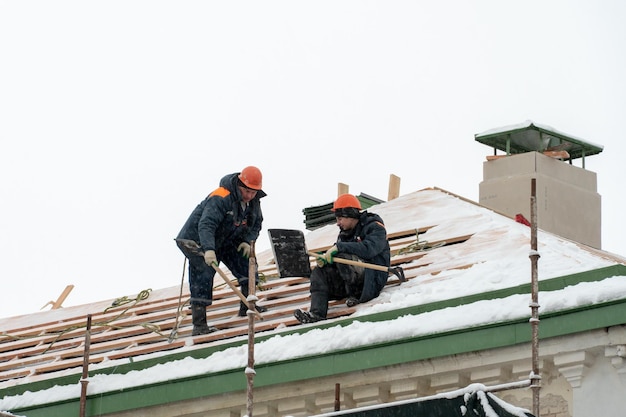 Dakreparaties in de winter Twee arbeiders repareren het dak op een ijzige en besneeuwde dag Mannen met schoppen verwijderen sneeuw van het dak