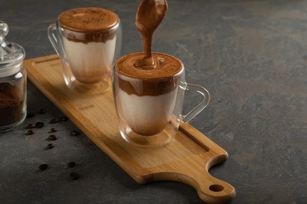 Фото Кофе dalgona, модный напиток. два стакана кофе dalgona на деревянной доске, рядом с кофейными зернами и растворимым кофе в стеклянной банке. вертикальная ориентация, с копией пространства.