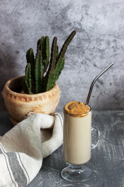 Dalgona Coffee in a transparent cup with a cocktail straw and cactus on a concrete table.