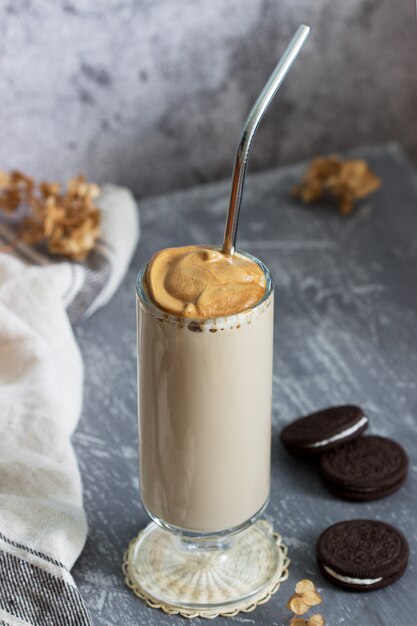 Caffè dalgona in una tazza trasparente con una cannuccia cocktail, biscotti e fiori secchi su un tavolo di cemento.