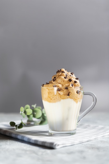 Dalgona schuimende koffie in een transparante glazen beker met chocoladedruppels, trendy Koreaanse drank gemaakt met melk en koffieschuim op grijze muur