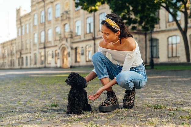 dame in stijlvolle vrijetijdskleding speelt met een schattige zwarte hond op straat met een glimlach op haar gezicht