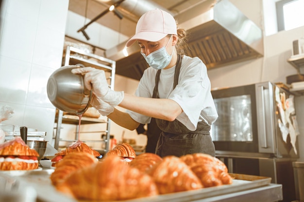 Dame met masker giet roze glazuur op croissant bij aanrecht in bakkerij