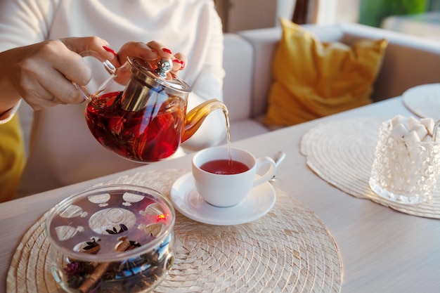 Dameshanden lichte huid met rode manicure giet thee uit een transparante theepot in een witte kop. Lichte levensstijlfoto van het ontbijt in een stijlvol café.