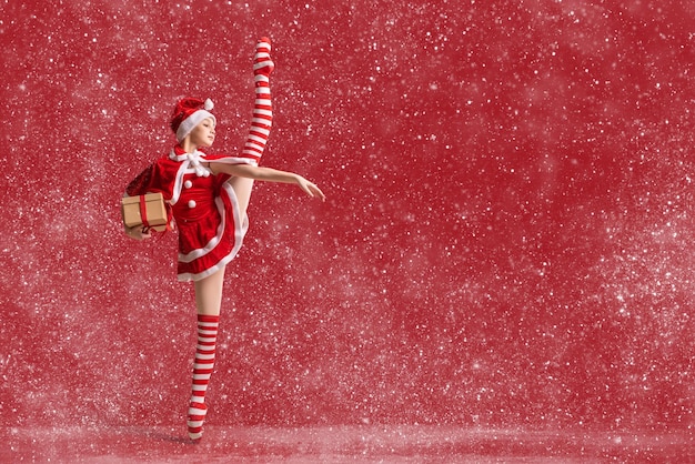 Танцующая балерина в пуантах с подарком в руках, одетая как Санта-Клаус на красном фоне