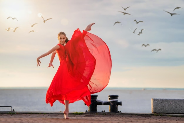 Dancing ballerina in a red flying skirt and leotard on the ocean embankment or on the sea beach surrounded by seagulls in the sky