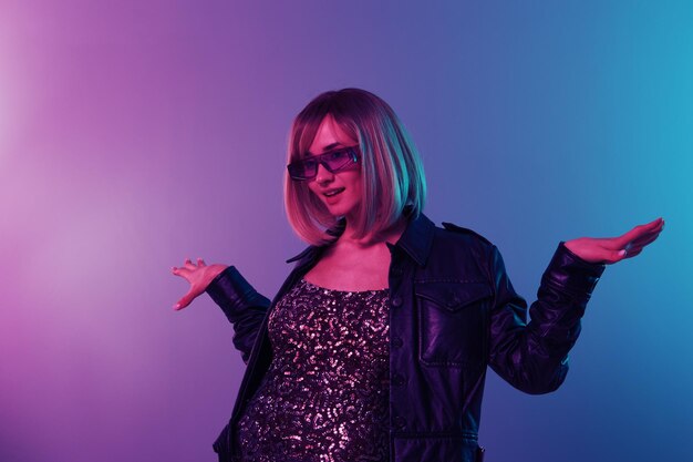 Dancing sexy beautiful blonde woman in leather jacket sparkly\
dress trendy sunglasses spread hands posing isolated in blue pink\
color light studio background neon party cyberpunk concept copy\
space