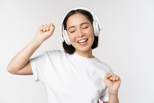 Ballare e cantare musica d'ascolto donna asiatica in cuffie in piedi in auricolari su sfondo bianco