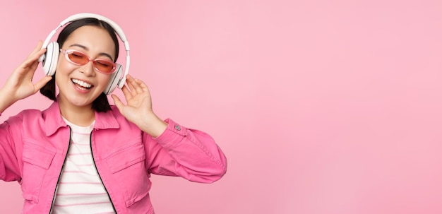 Dancing stylish asian girl listening music in headphones posing against pink background