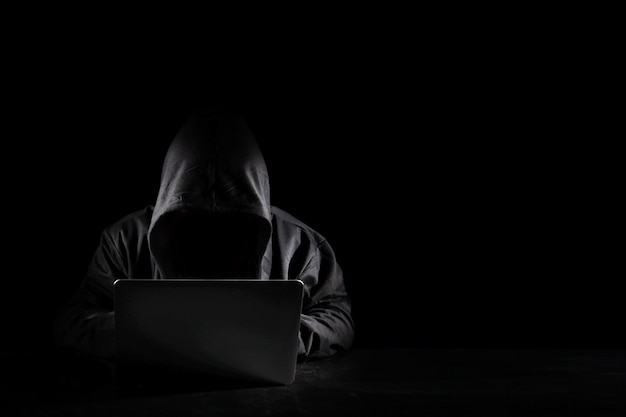 Dangerous anonymous hacker man in black hooded using computer, breaking into security data corporate server. He sitting, working on black background. Internet crime, cyber attack security concept