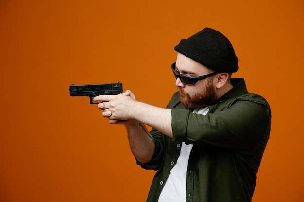 Dangerous looking bearded man wearing black glasses and hat holding a gun looking aside standing over orange background