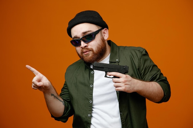 Dangerous looking bearded man wearing black glasses and hat holding a gun pointing with index finger to the side standing over orange background