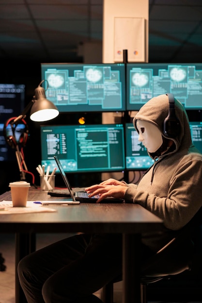 Dangerous masked hacker stealing information from database server online, hacking computer hardware system. Impostor with hood and mask creating malware and security breach in office.