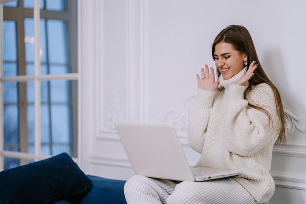 Foto dankbare spaanse jonge vrouw in witte trui die thuis zit met laptop maakt videogesprek