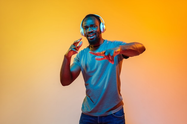 Dansen, blij. Portret van een Afro-Amerikaanse man in een koptelefoon op een oranje achtergrond met kleurovergang in neonlicht. Mooi afromodel. Concept van menselijke emoties, gezichtsuitdrukking, verkoop, advertentie. Kopieerruimte.