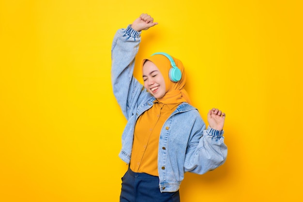 Dansende jonge Aziatische vrouw in een spijkerjasje met een koptelefoon om naar muziek te luisteren, geïsoleerd op een gele achtergrond