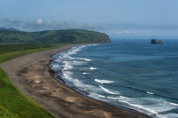 Dark almost black color sand beach of Pacific ocean. Stone mountains and yellow grass are on a background. Light blue sky.