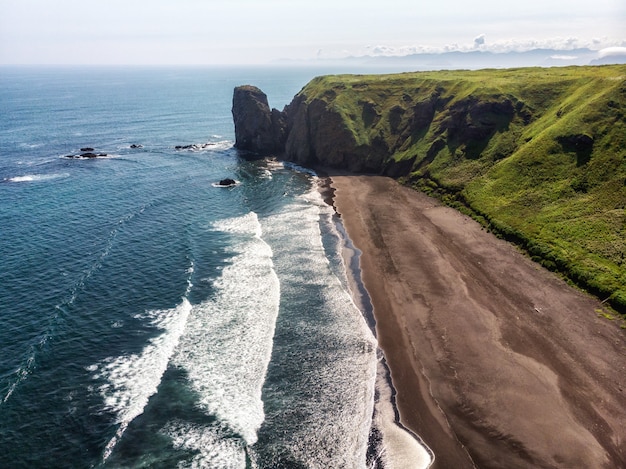 Dark almost black color sand beach of Pacific ocean. Stone mountains and yellow grass are on a background. Light blue sky.