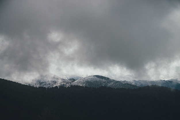 Dark atmospheric landscape with forest mountains in snow in low clouds in overcast weather. Minimalist gloomy scenery with gray rain clouds above snowy hills and mountain silhouettes. Grainy clouds.