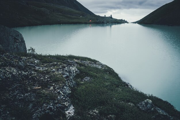 Dark atmospheric landscape with orange tent near mountain lake in highland valley under cloudy sky in dark time. Ripples on mountain lake water in dusk. Dark scenery with water ripple on highland lake