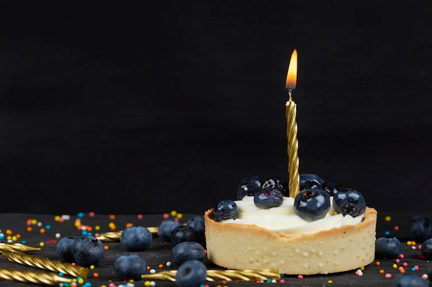 On a dark background a cake with a burning candle sprinkled with black berries