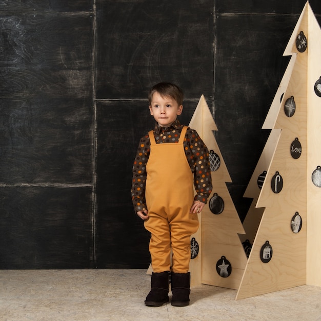 Su uno sfondo scuro un ragazzino in posa con un costume alla moda vicino a un albero di natale di legno