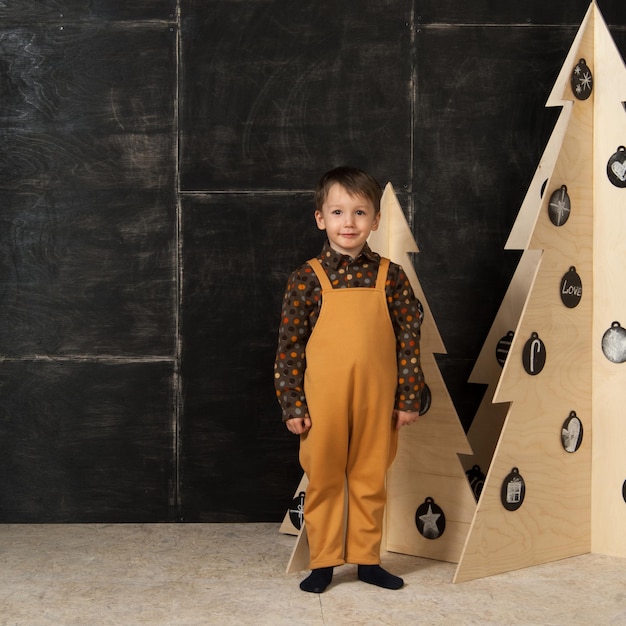 On a dark background little boy posing in a fashionable costume near a Christmas tree made of wood