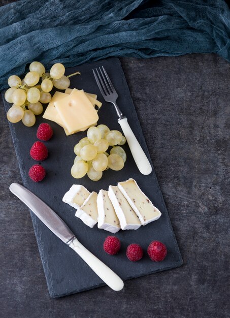 dark background and a stone board, on it raspberry, grapes and cheese 