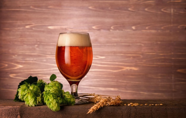 Фото Бокал темного пива с пеной и зелеными шишками хмеля на деревянном фоне с копией пространства