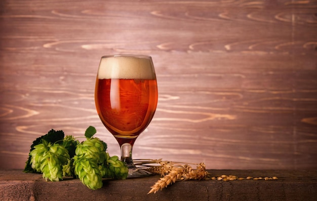 Dark beer glass with foam and green hop plant cones on wooden background with copy space