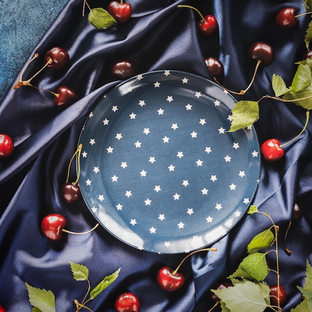 Темно-синяя композиция с пустой синей тарелкой со звездным узором и ягодами вишни с листьями.